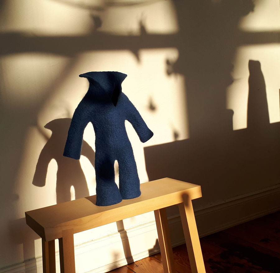 Das Foto zeigt die aus Wolle gefilzte Skulptur eines Blaumanns, der auf einem Holzhocker vor einer Wand steht. Die Wand zeigt vielfältige Schatten